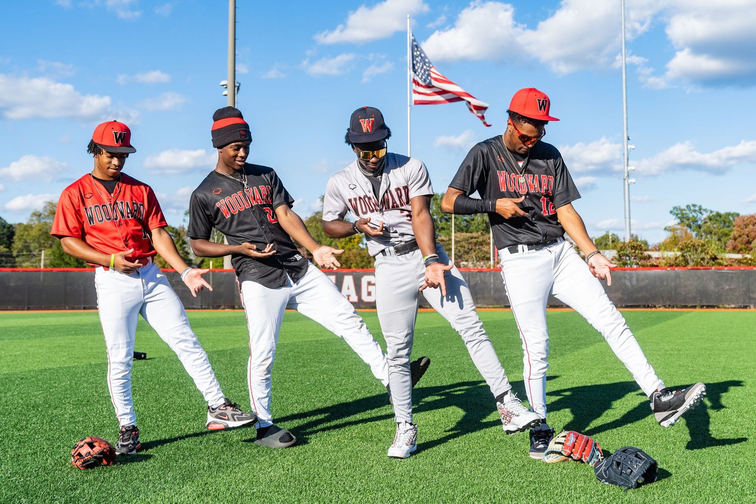 Four baseball players in red and black uniforms stretch and warm up on a green field with an American flag in the background.