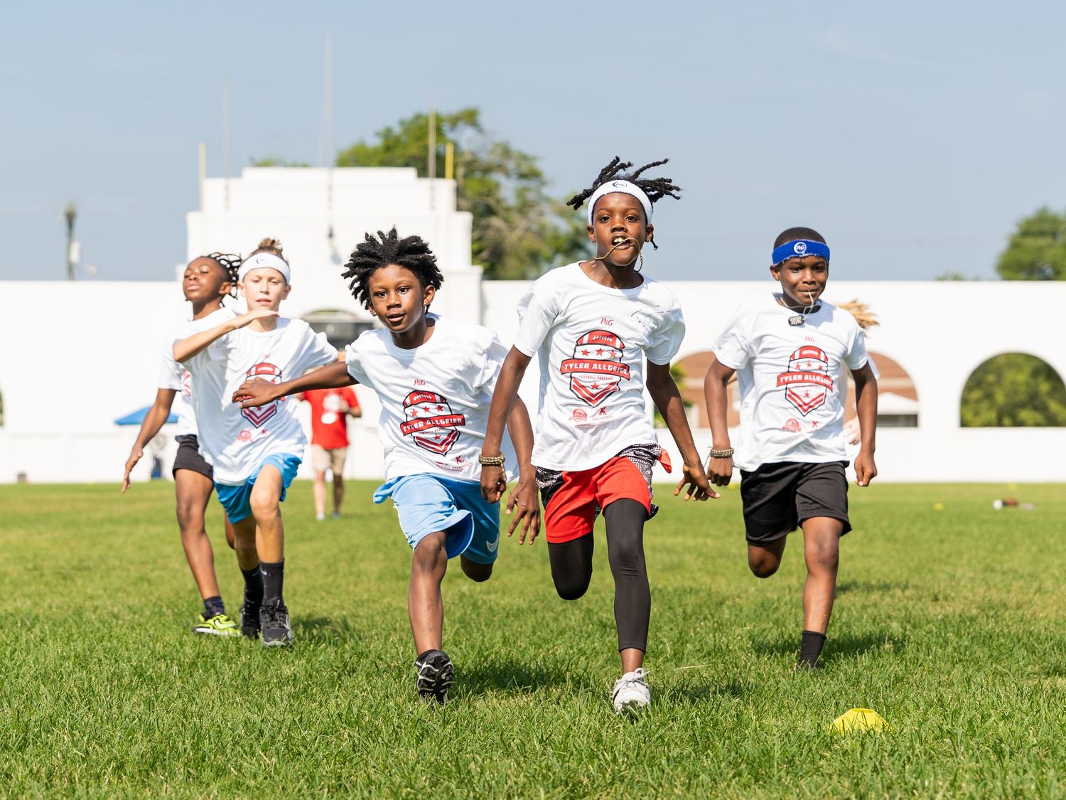 Kids in white camp shirts racing across a green field during an outdoor youth football sports camp event.