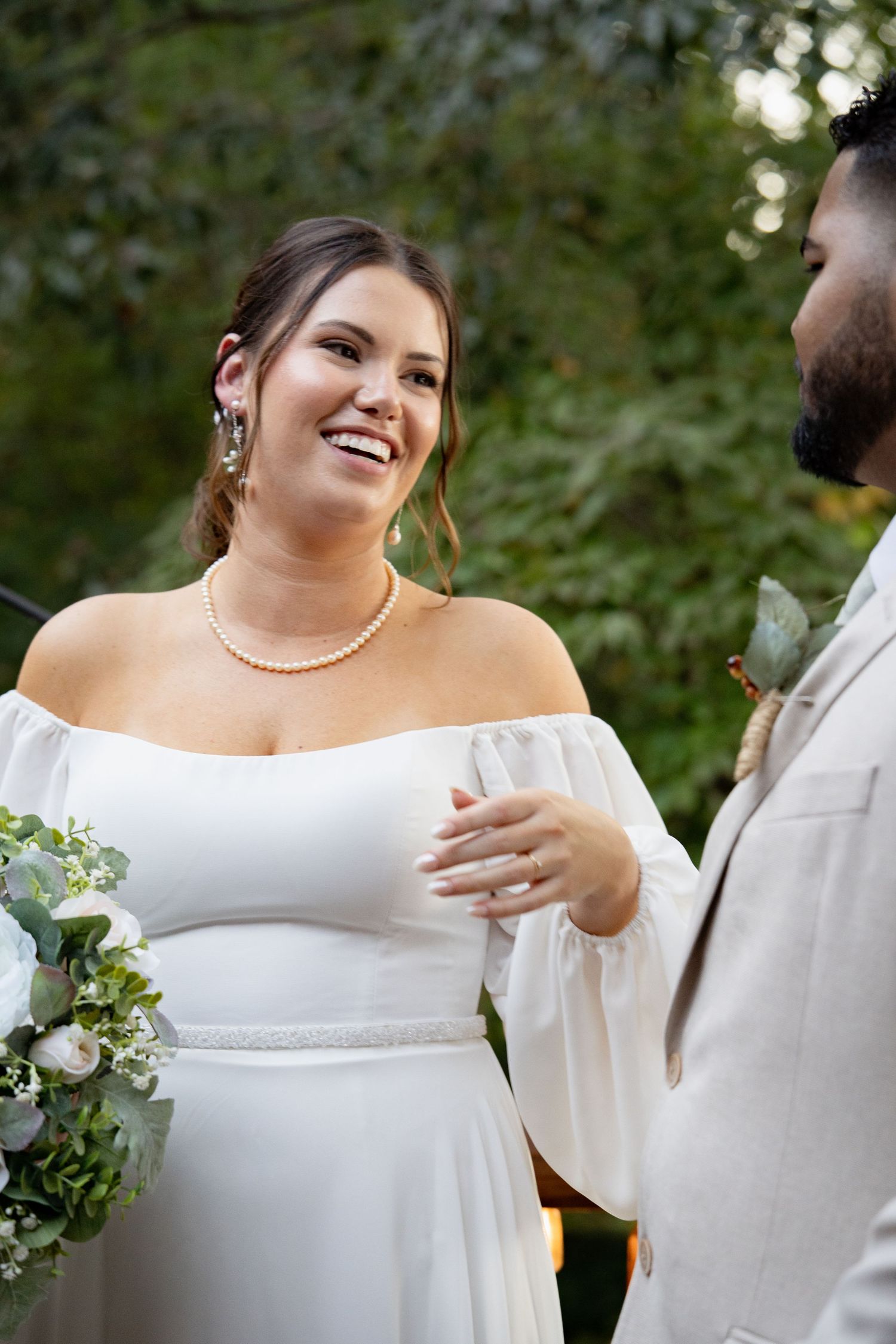 Smiling bride in white off-shoulder gown with pearl necklace holds bouquet during outdoor wedding ceremony.