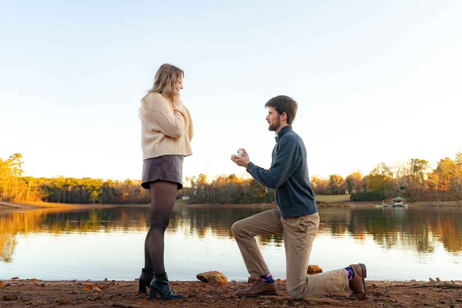 Man proposes to woman on one knee by a lake during autumn sunset, holding a ring box in a romantic outdoor setting.