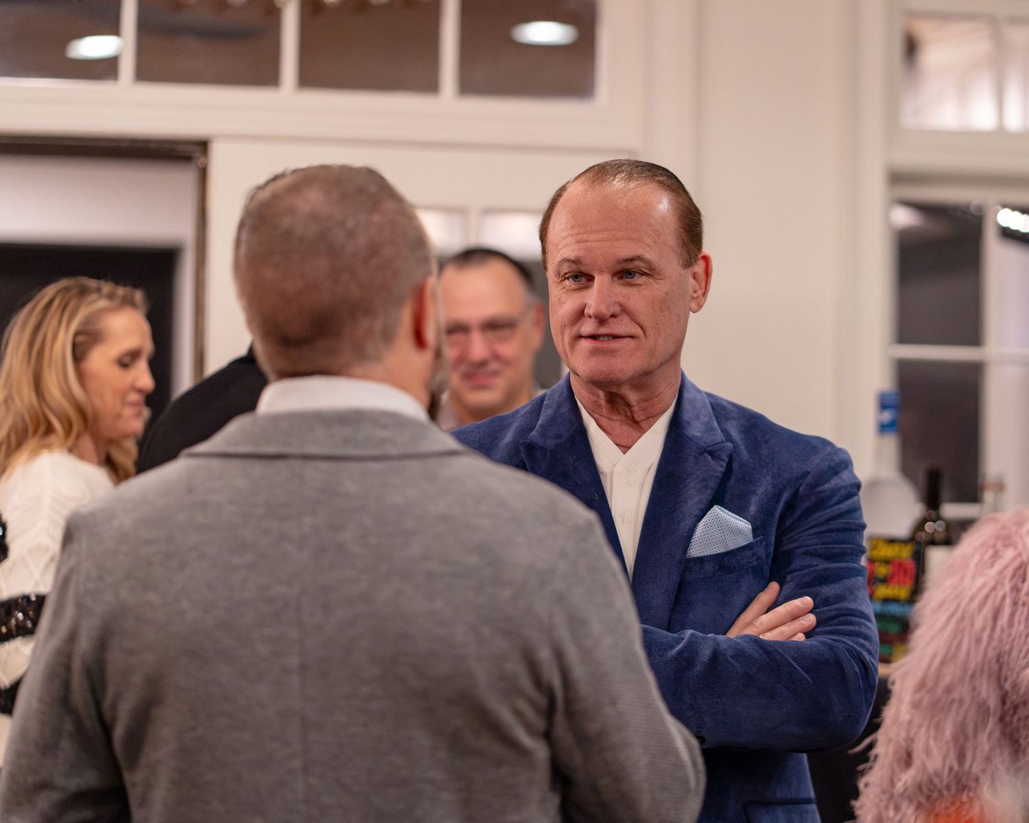 Man in blue blazer with arms crossed networking at an indoor social gathering event.