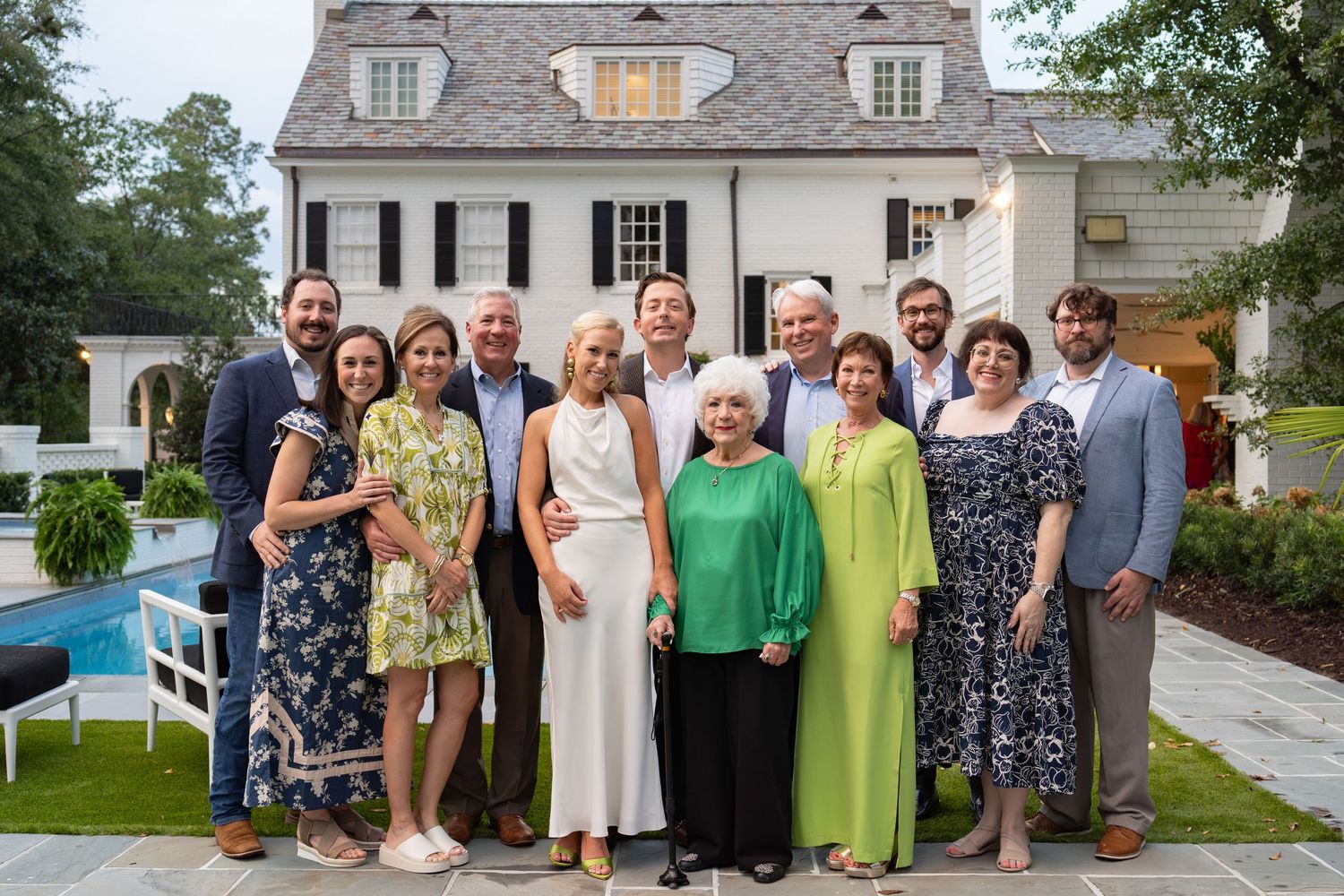 Large family group posing outdoors in front of elegant white colonial home with pool, dressed in summer formal attire.