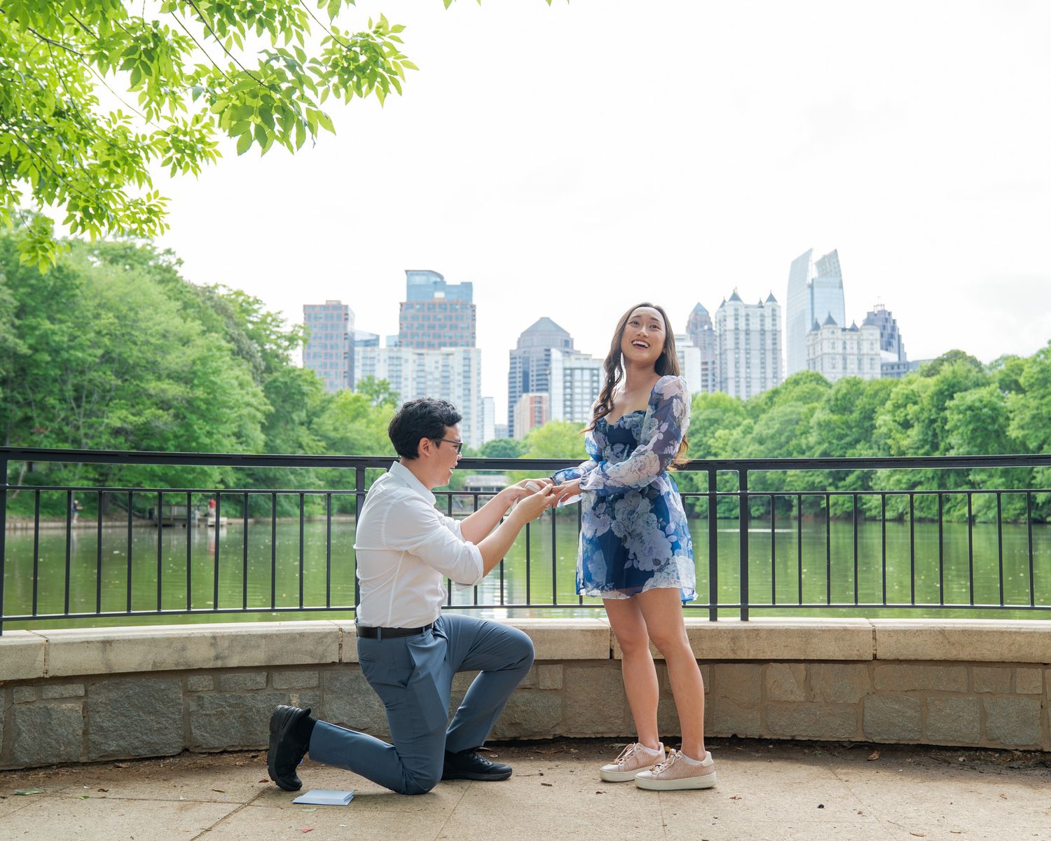 Man proposes on one knee to woman in floral dress at park with city skyline backdrop.