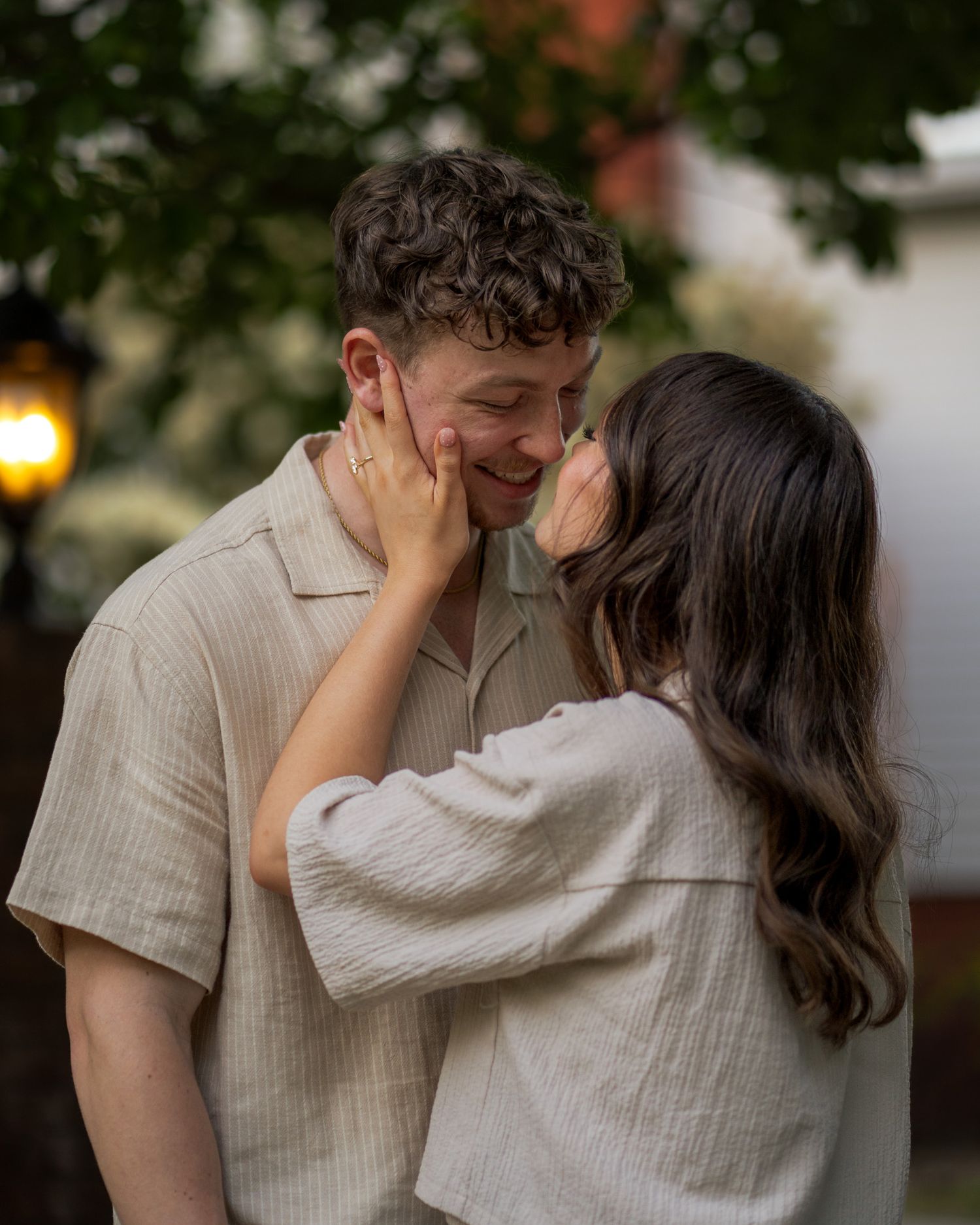 A couple in matching beige outfits share a tender moment outdoors, with the woman gently cupping the mans smiling face.