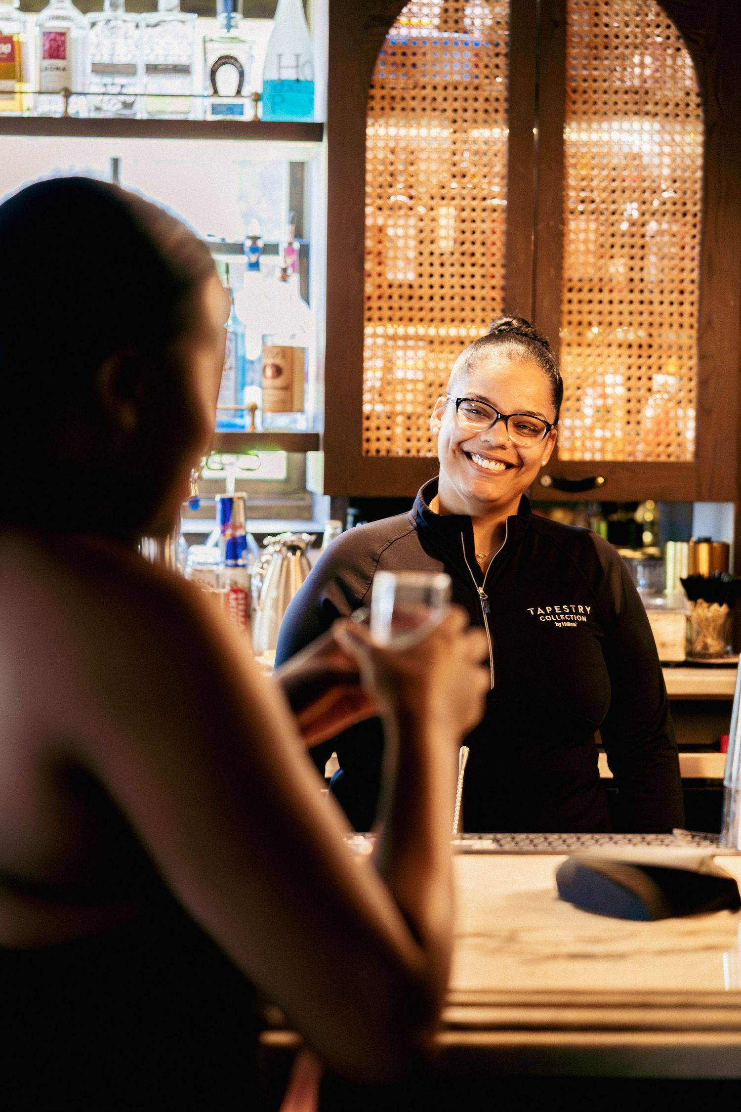 Smiling bartender in Tapestry uniform serves a customer at a warmly lit bar with shelves of bottles.