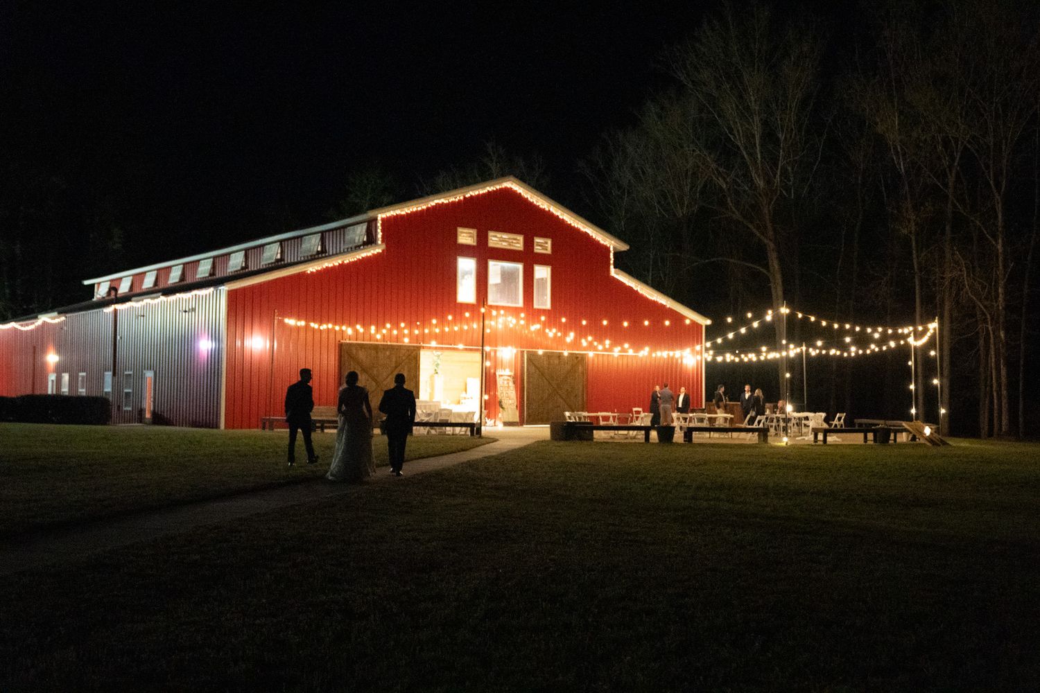 Red barn venue lit up at night with string lights and guests gathered outside on a green lawn.
