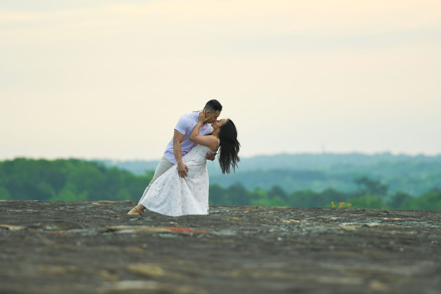 A romantic couple embraces during a sunset photoshoot on a hilltop overlooking a scenic landscape.