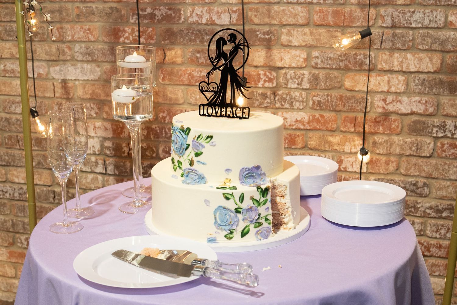 Two-tier white wedding cake with floral designs and couple silhouette topper on a lavender tablecloth.