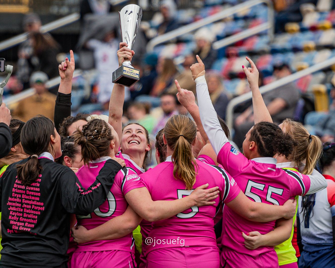 Women's soccer team in pink jerseys celebrating championship victory, raising trophy together on field.
