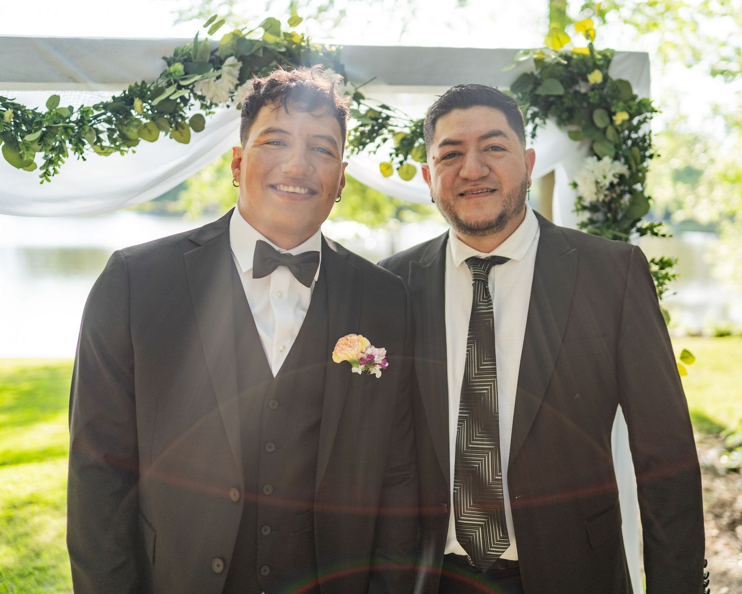 Two men in dark suits smile together outdoors at a wedding ceremony beneath a floral arch.