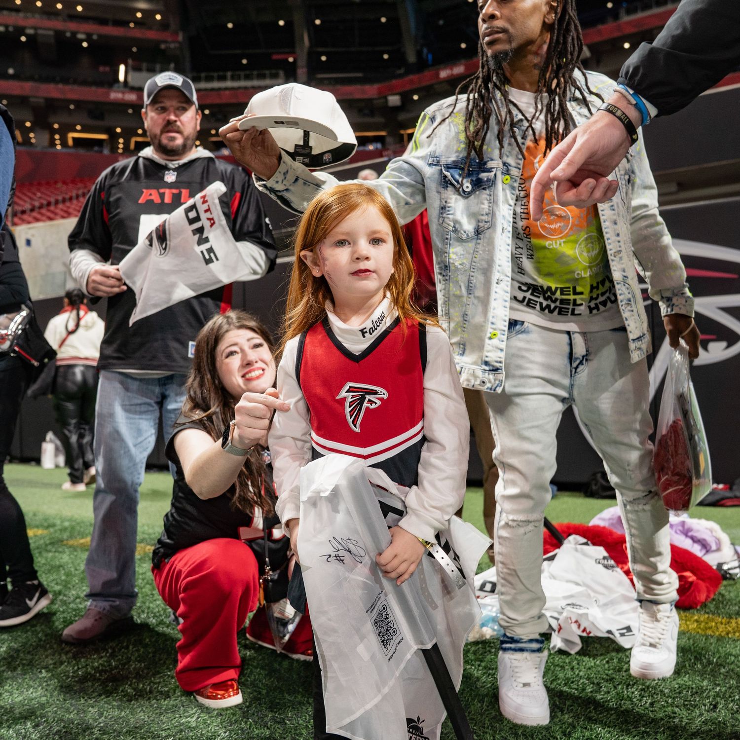Young girl in Atlanta Falcons cheerleader uniform stands on stadium field surrounded by fans and players.