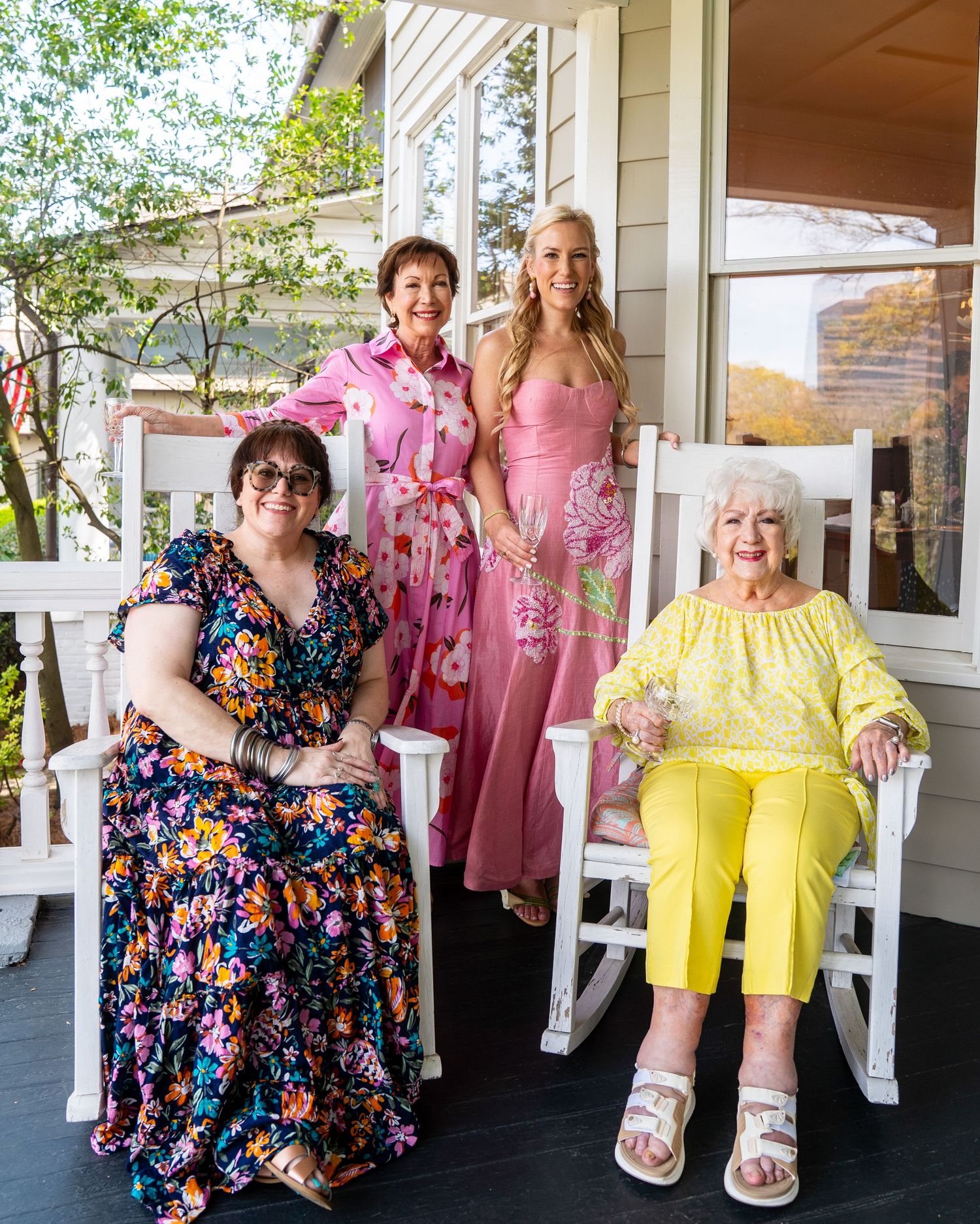 Four women in colorful spring dresses posing on a white rocking chair porch for a portrait.