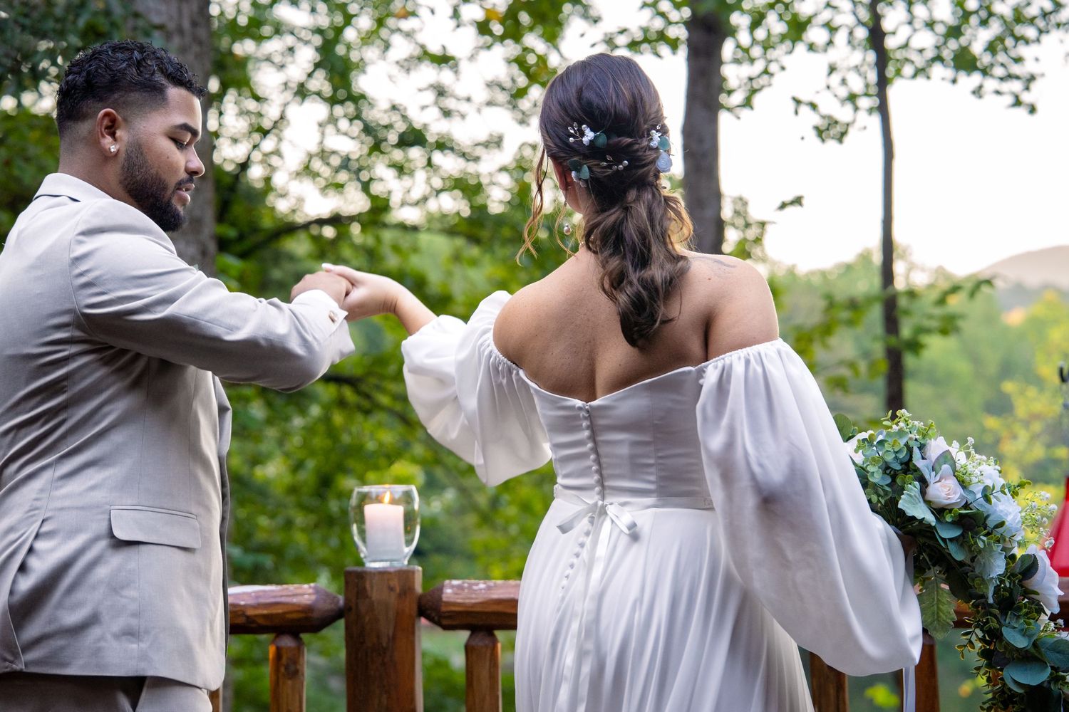 Bride and groom hold hands during outdoor wedding ceremony on wooden deck surrounded by lush green trees.