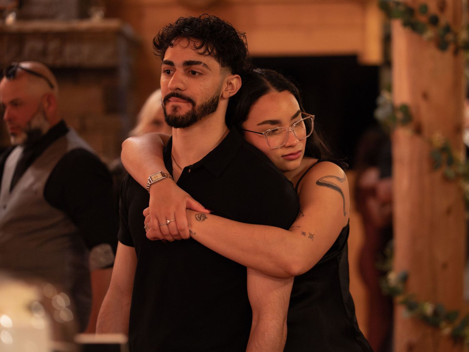 Couple embracing at a wedding reception in a rustic venue, both wearing black outfits.