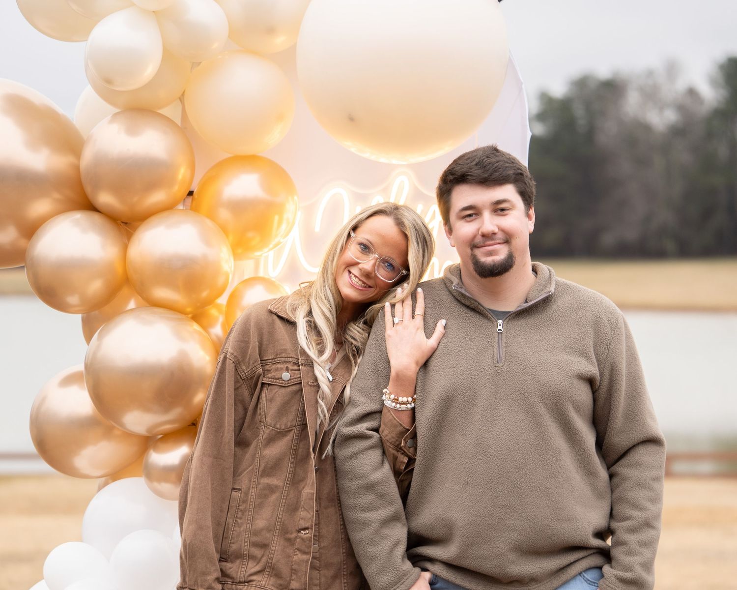 Couple smiling in front of gold and white balloon arch, woman showing off engagement ring outdoors.