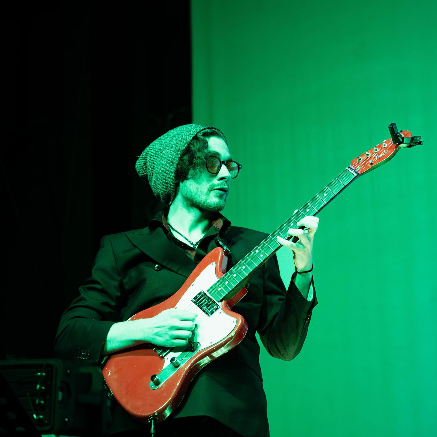 A musician wearing a green beanie and glasses plays a red electric guitar on a green-lit stage.