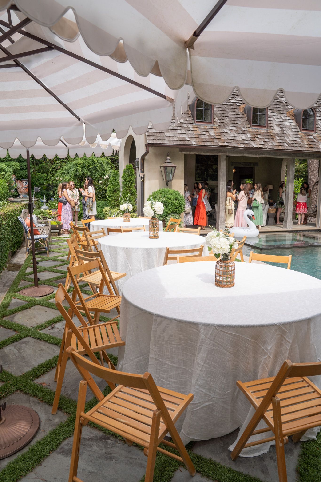 Elegant outdoor wedding reception with round white-draped tables, wooden folding chairs, striped tent, and guests near a pool.