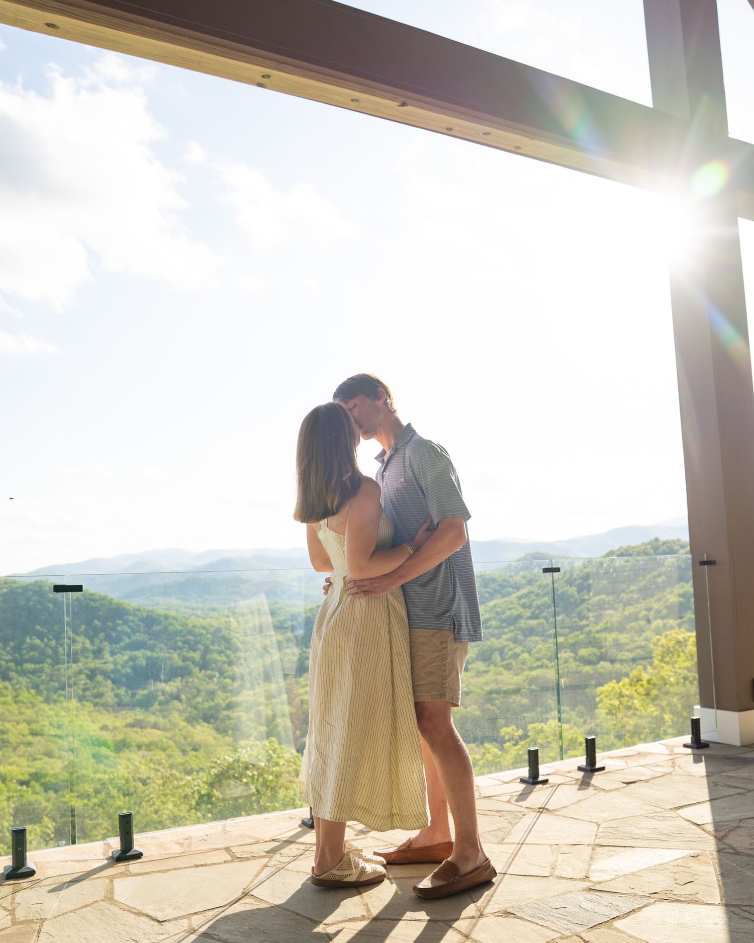 Couple kissing on a sunlit terrace overlooking lush green hills, with golden sunlight streaming through the open structure.