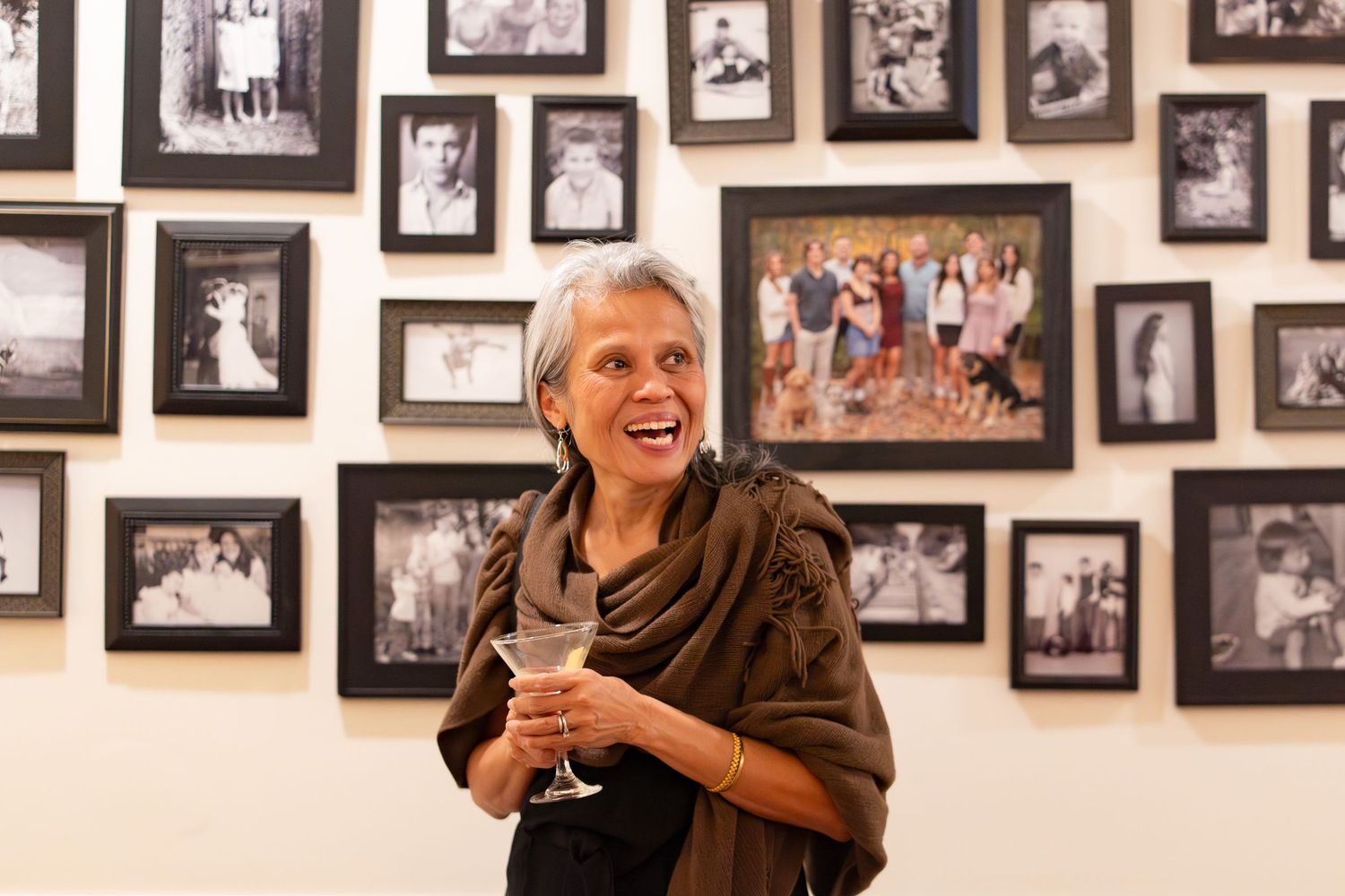 Smiling woman holding a wine glass standing in front of a wall of framed family photos at a gathering.