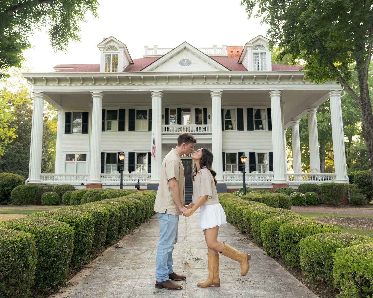 Couple shares romantic kiss in front of grand white Southern plantation mansion with tall columns and manicured hedges.