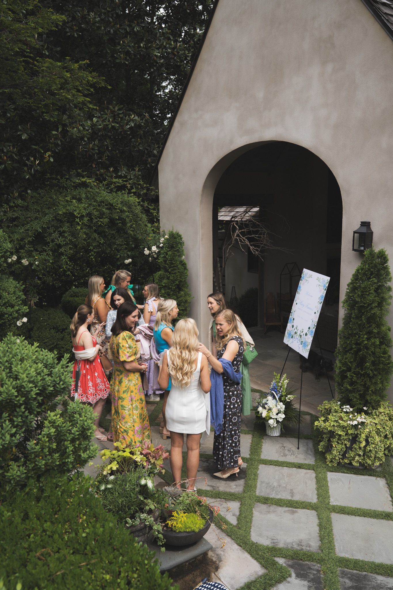 Group of women in colorful dresses gathered outside an elegant stucco home entrance with lush garden landscaping.