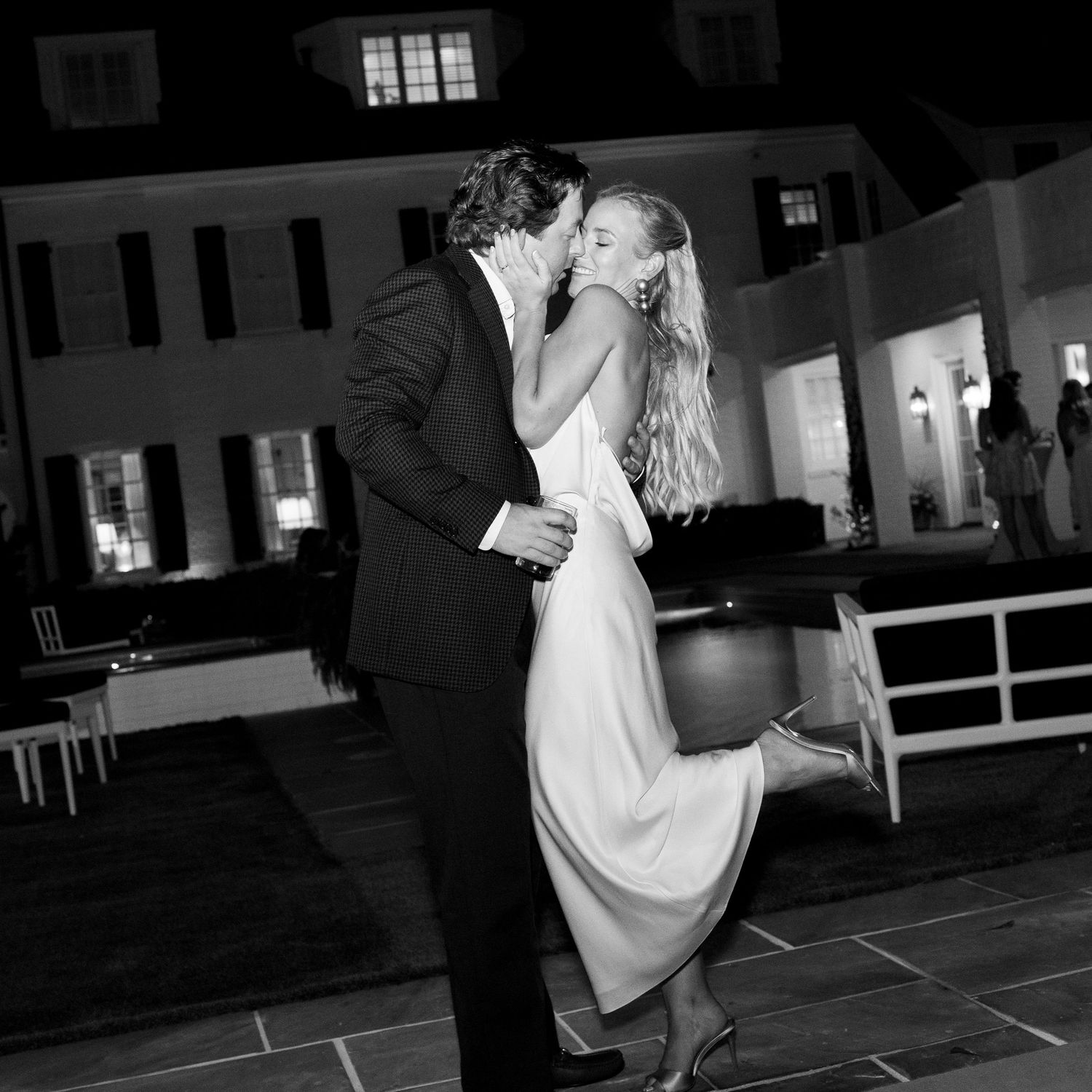Black and white photo of a couple sharing a romantic kiss at an outdoor wedding reception at night.
