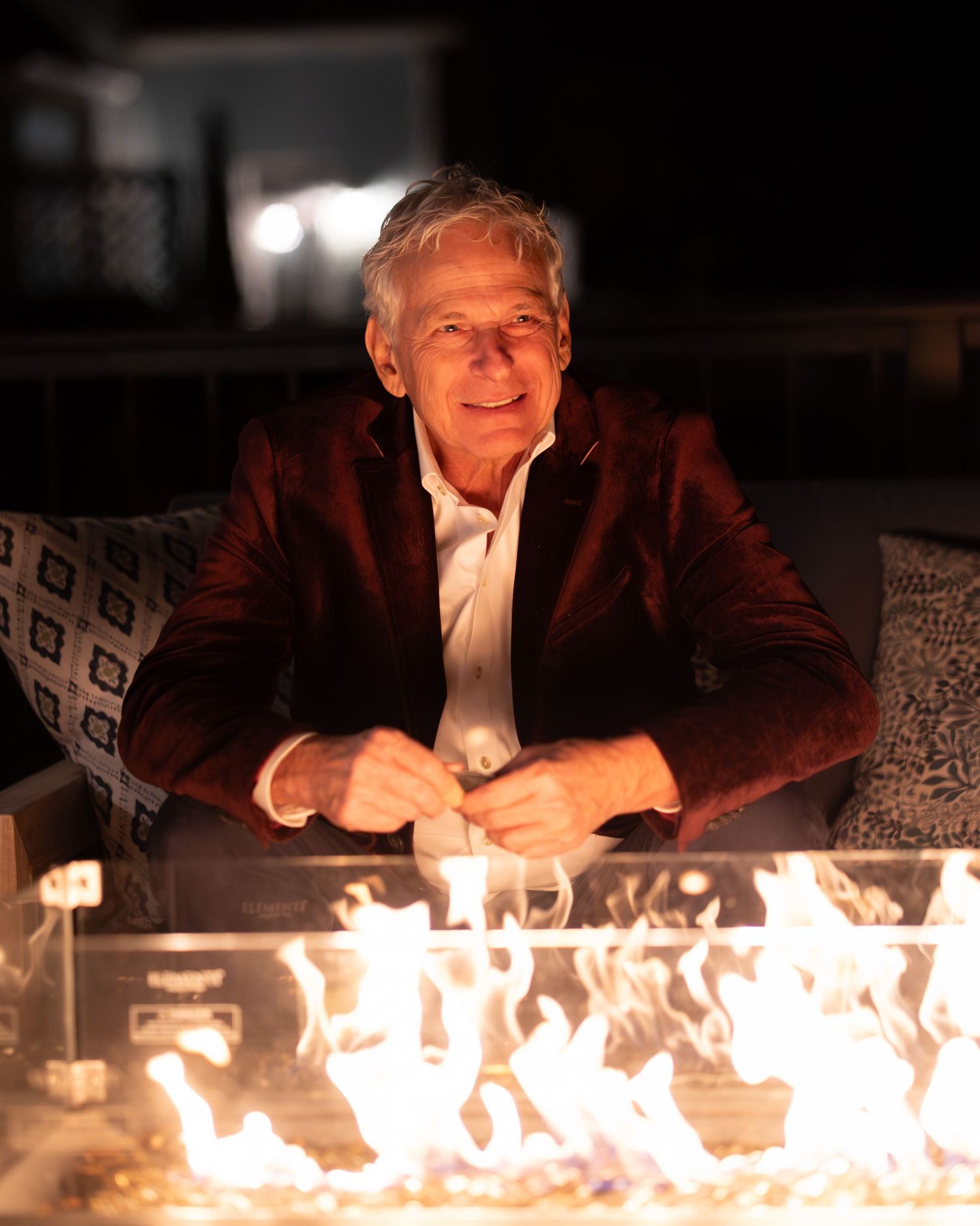 Elderly man in burgundy velvet blazer sitting by an outdoor fire pit at night.