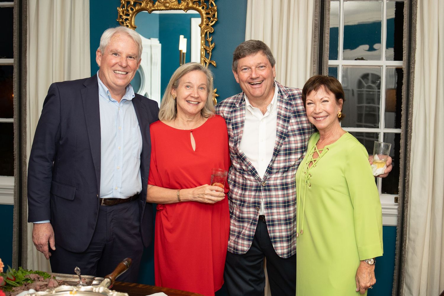 Two couples posing indoors at a social gathering, dressed in elegant evening attire with wine glasses.