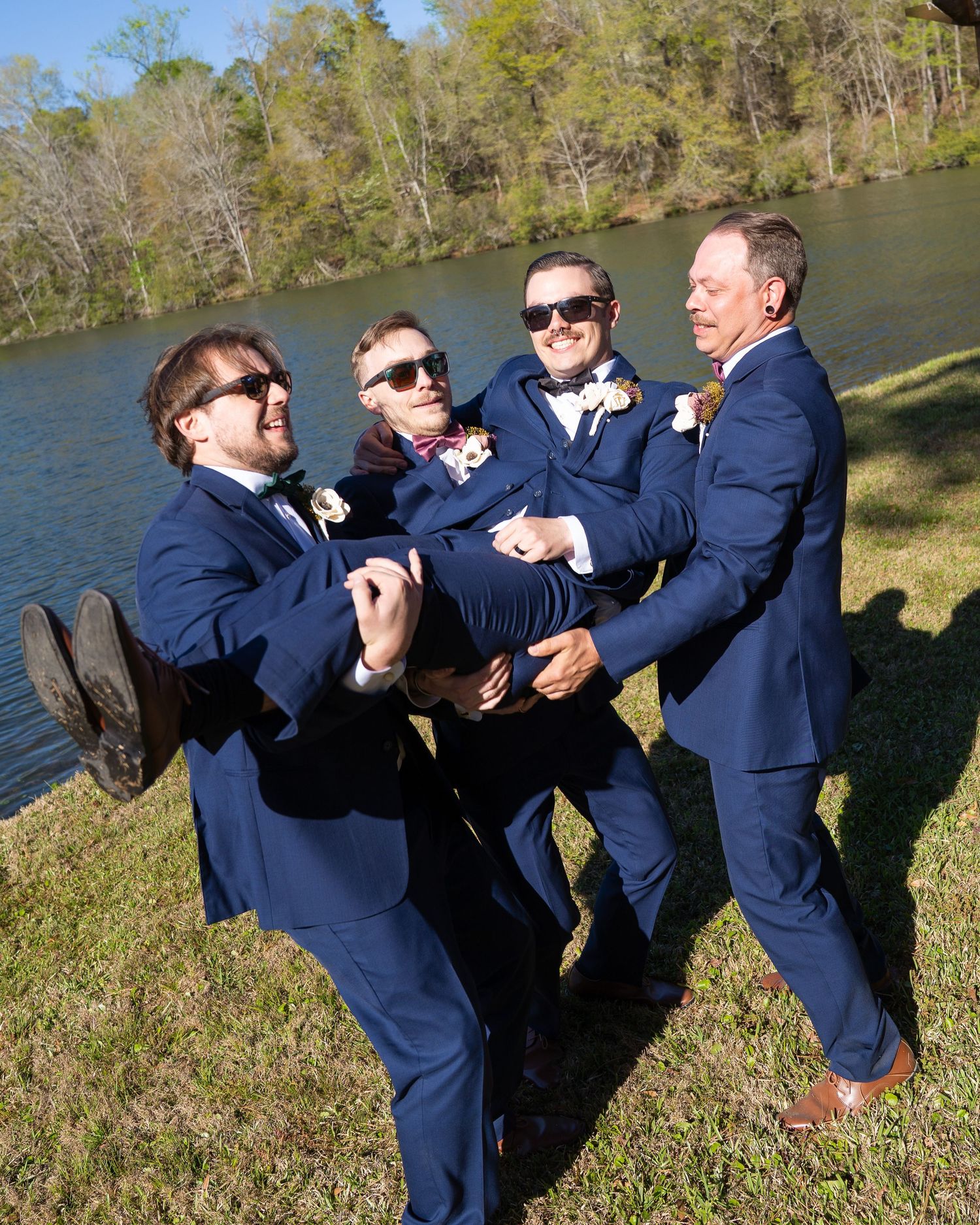 Groomsmen in navy blue suits and sunglasses playfully carry the groom outdoors near a scenic lake.