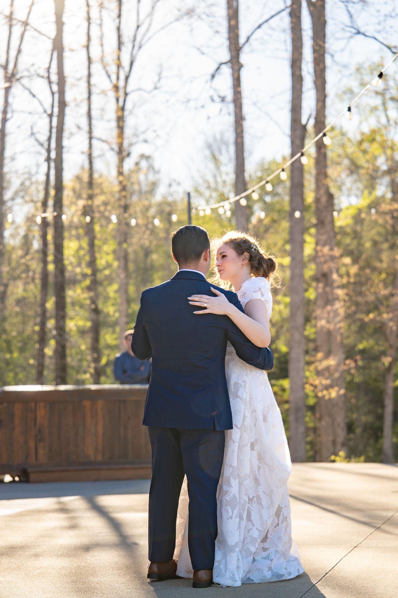 A bride and groom share their first dance outdoors surrounded by tall trees and string lights on a sunny day.