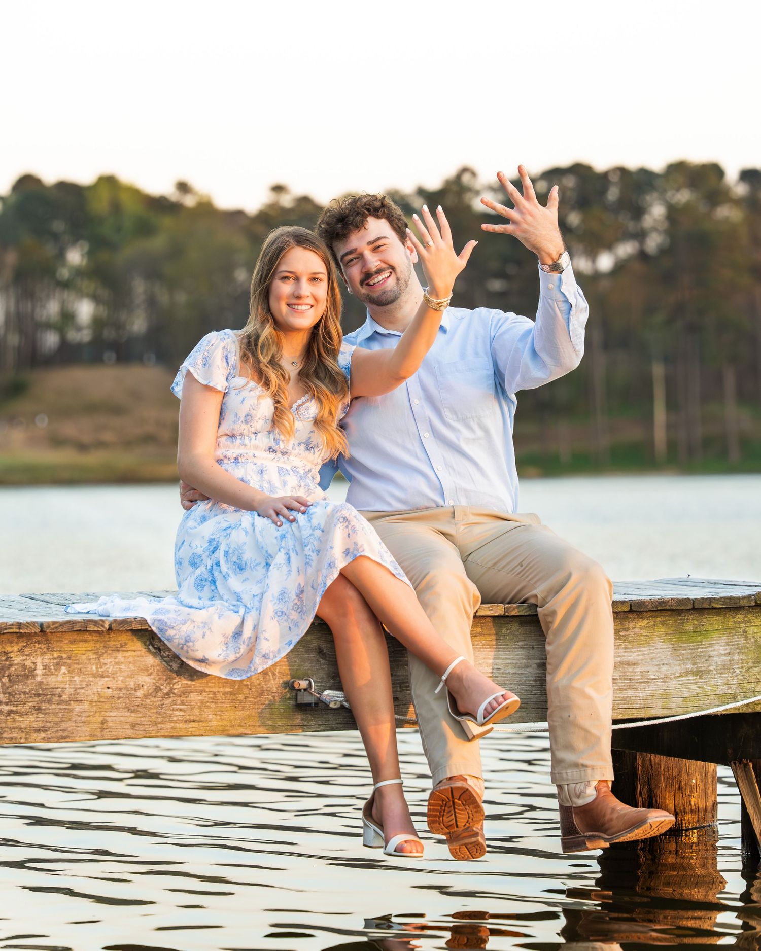 Happy couple sitting on a wooden dock over a lake, showing off an engagement ring at golden hour.
