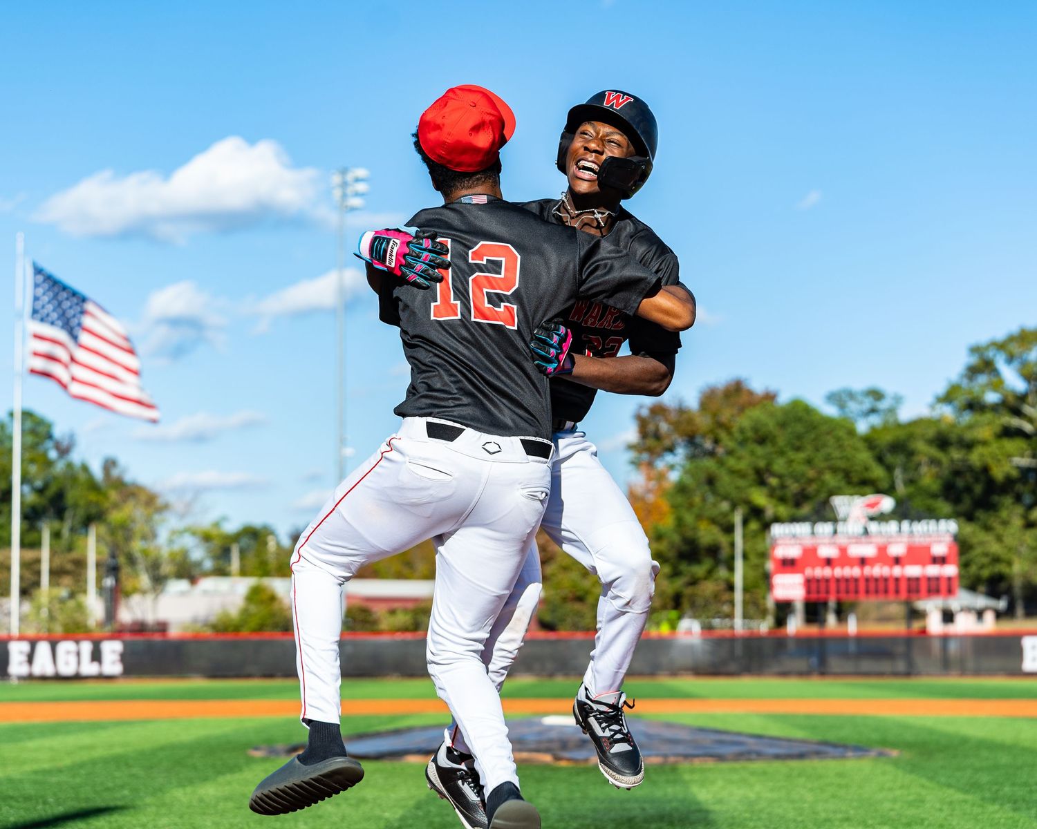 Two baseball players in black uniforms celebrate with a jump hug on the field near an American flag.