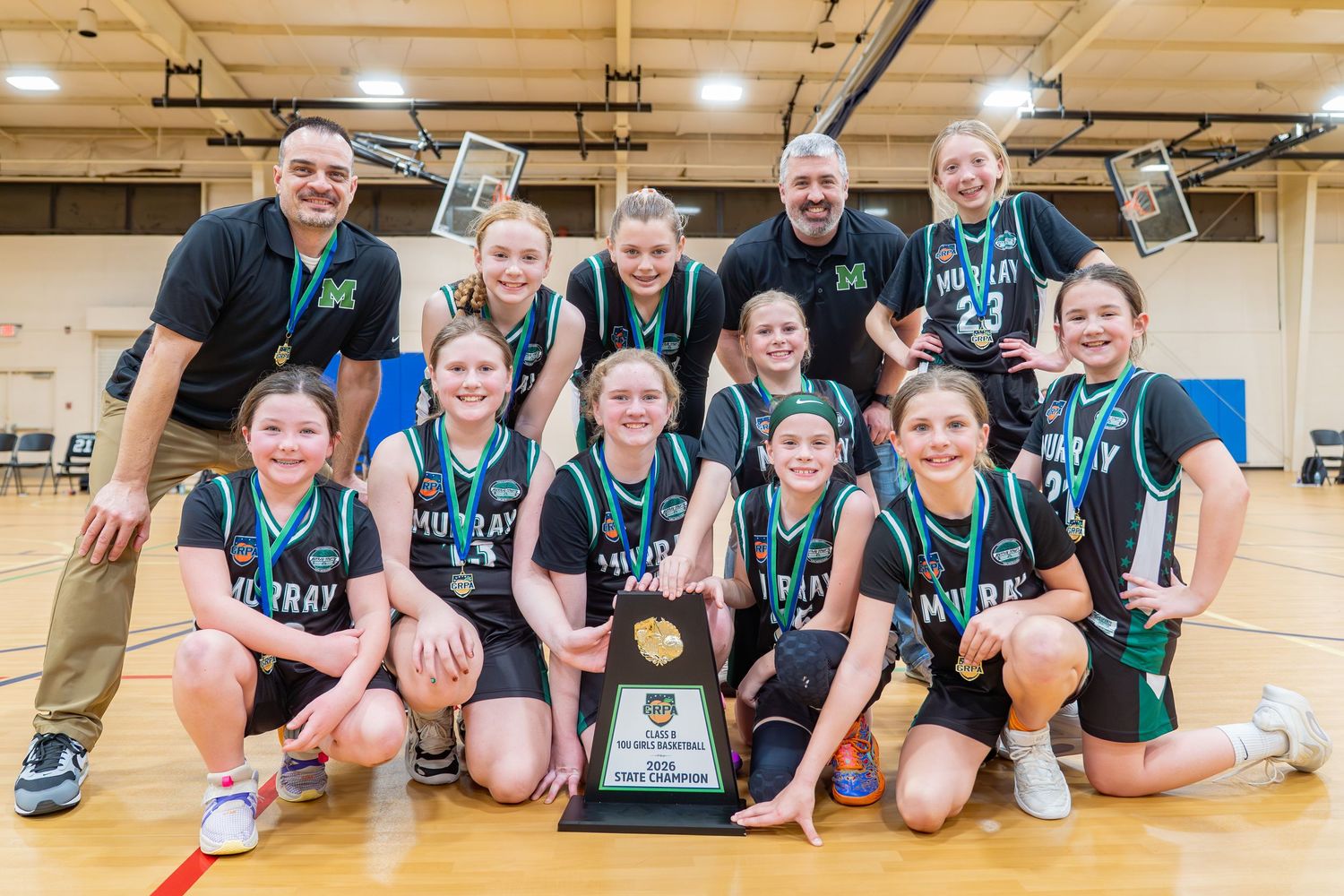 Youth girls basketball team poses with championship trophy on court, wearing teal uniforms with two adult coaches.