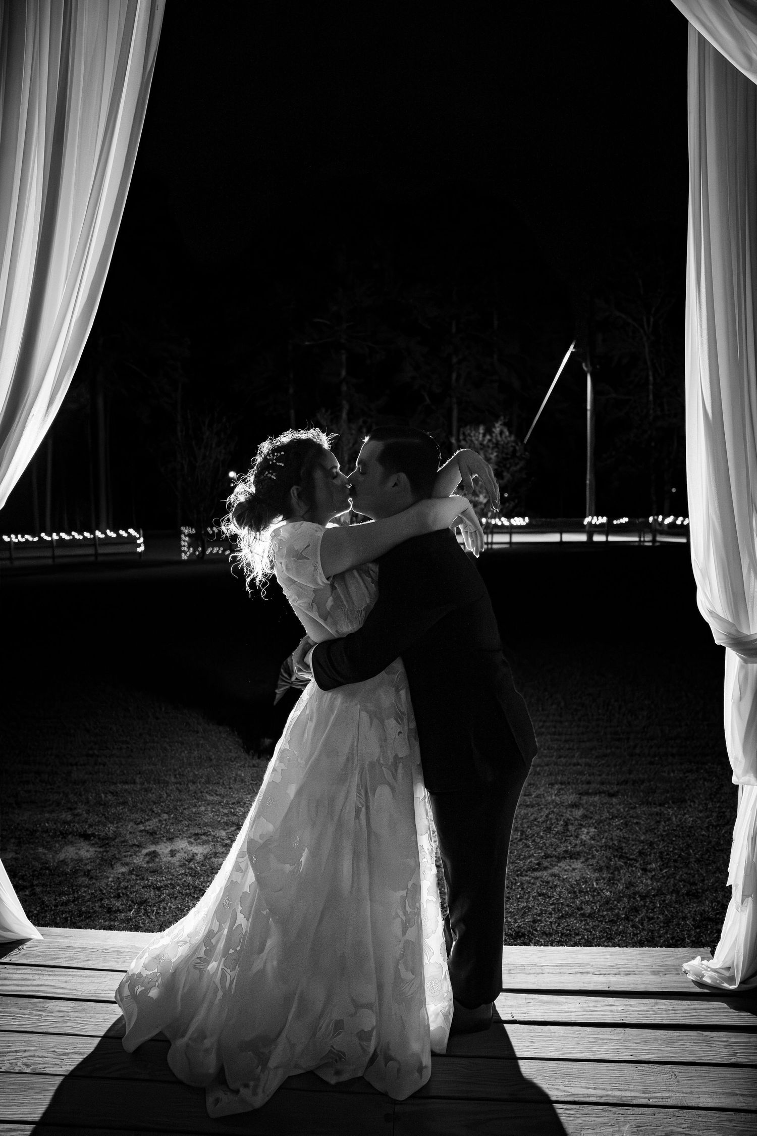 Bride and groom share their first dance on a wooden deck under white curtains at night in black and white photograph.
