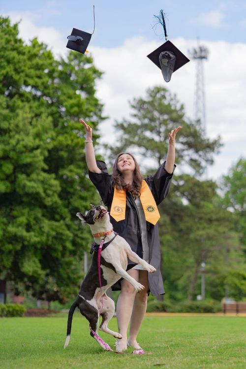 Graduate in cap and gown celebrates with jumping dog in sunny outdoor setting.