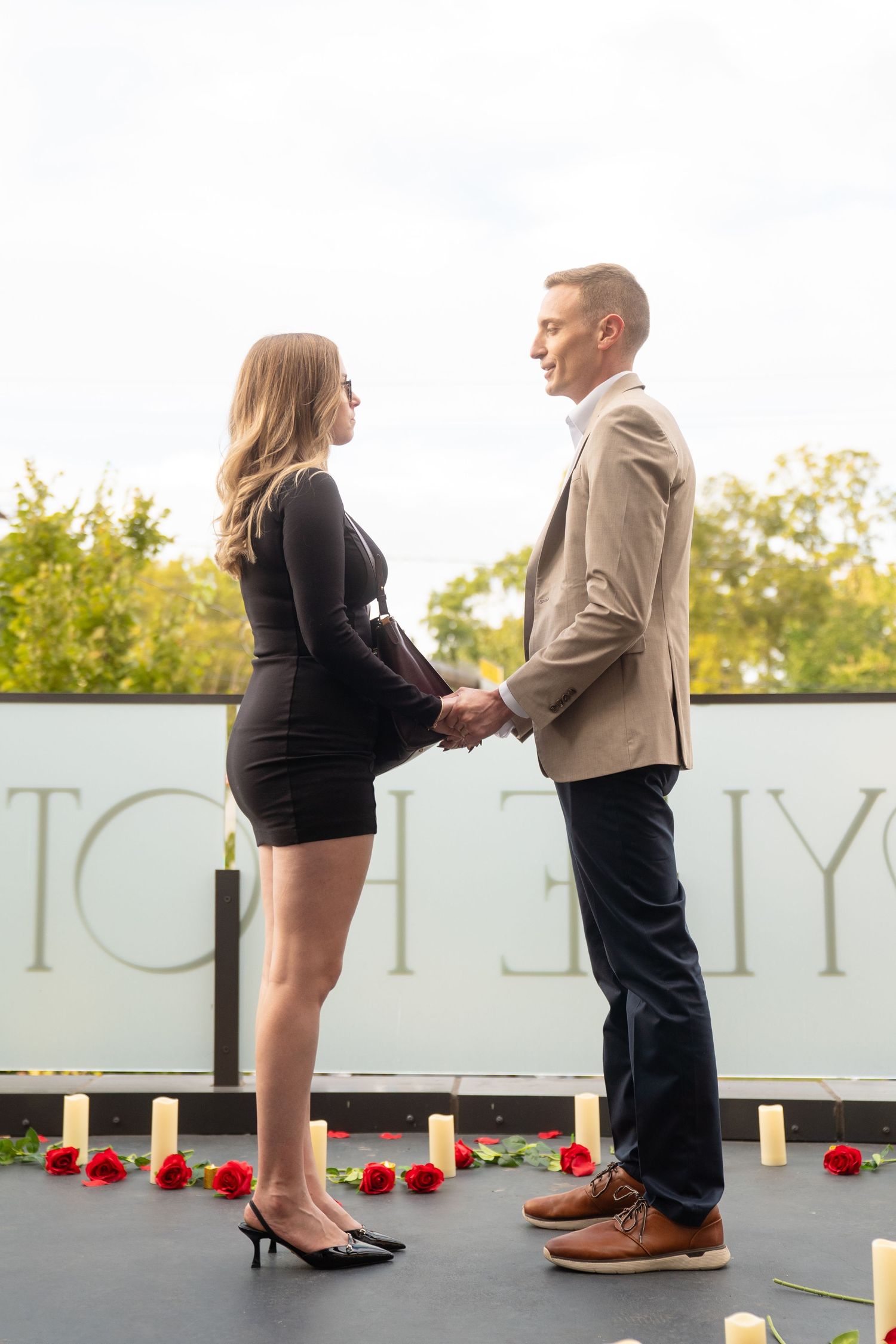 Romantic proposal scene on an outdoor terrace with red rose petals and candles.