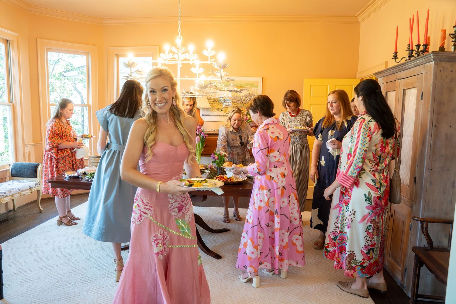 Woman in pink dress smiling at a colorful indoor gathering in a yellow-walled room with chandelier.