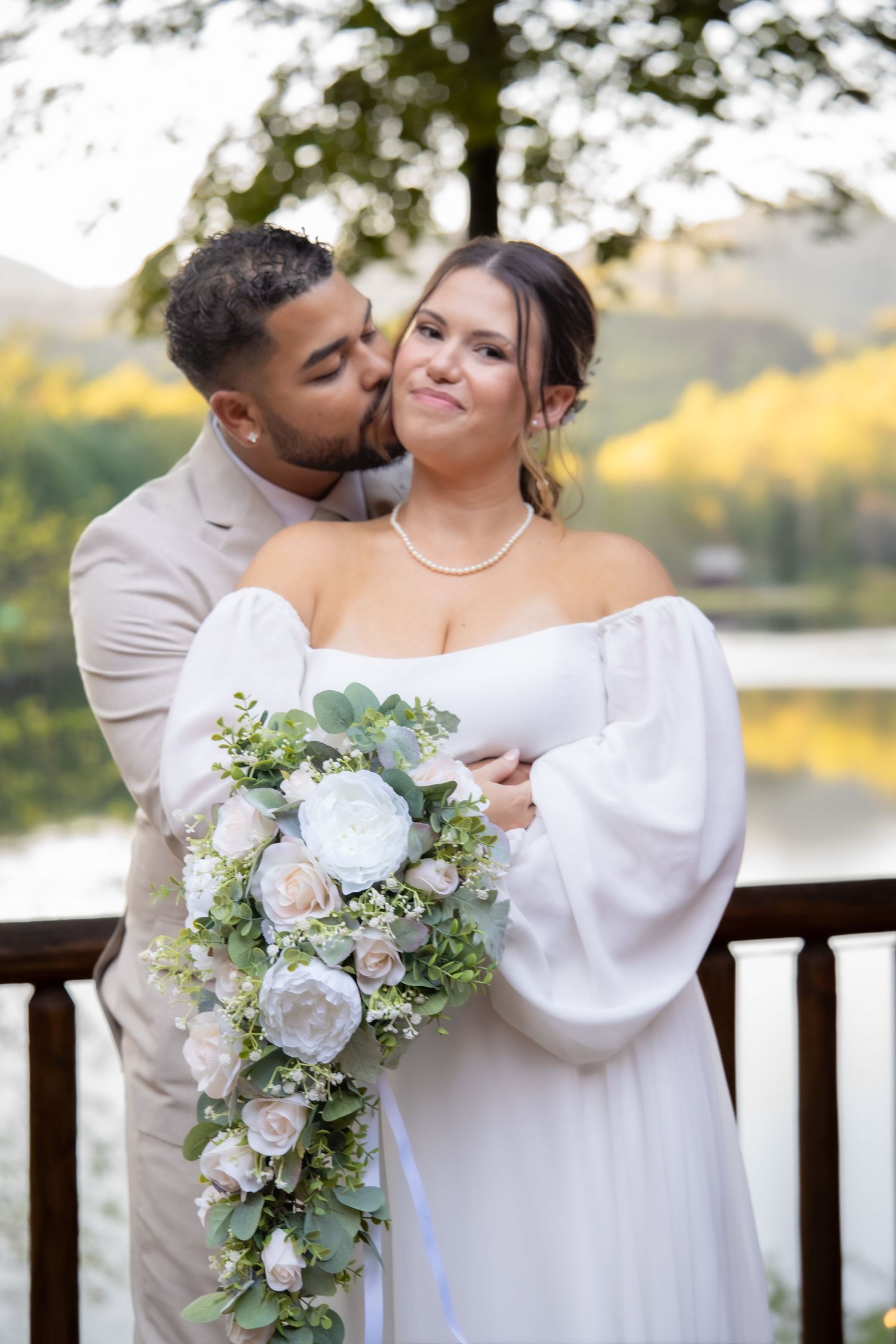 Groom in tan suit kisses bride in white off-shoulder gown holding cascading floral bouquet outdoors.