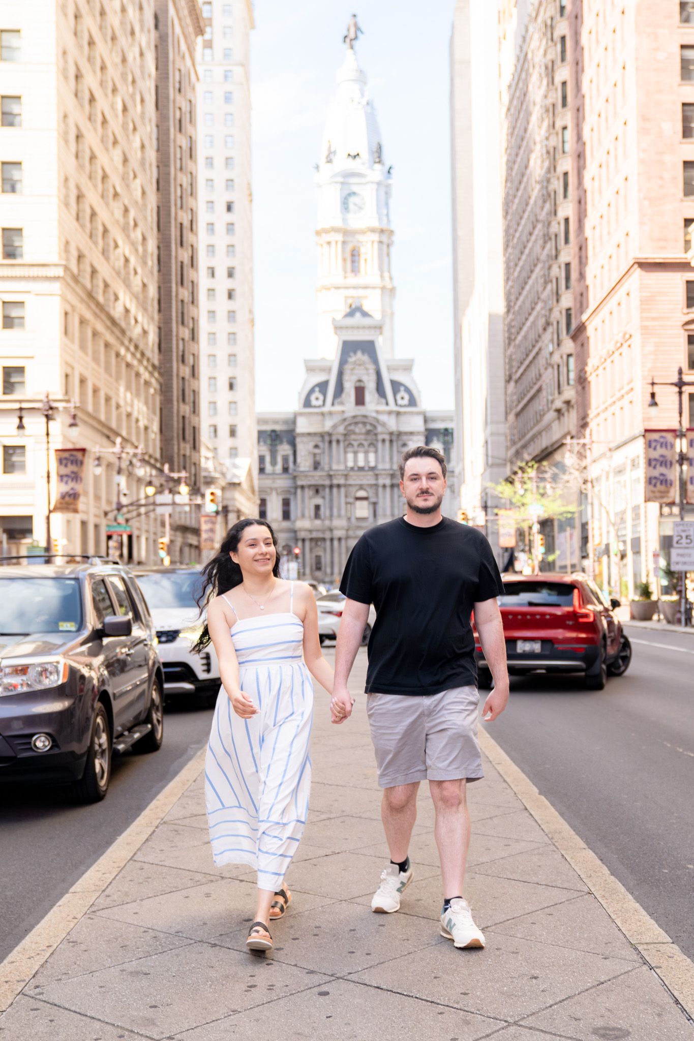 Couple holding hands walking on a city street with Philadelphia City Hall in the background on a sunny day.