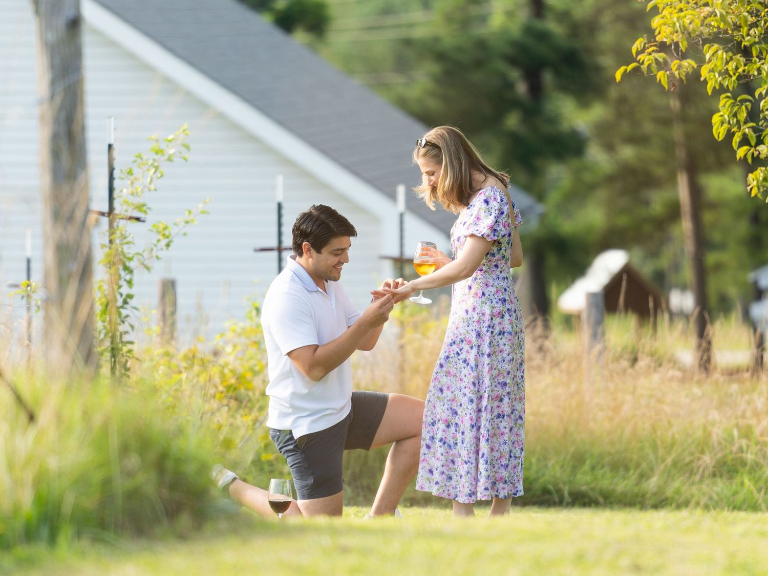 Man proposing to woman outdoors on sunny day, kneeling with ring near white chapel and lush greenery.