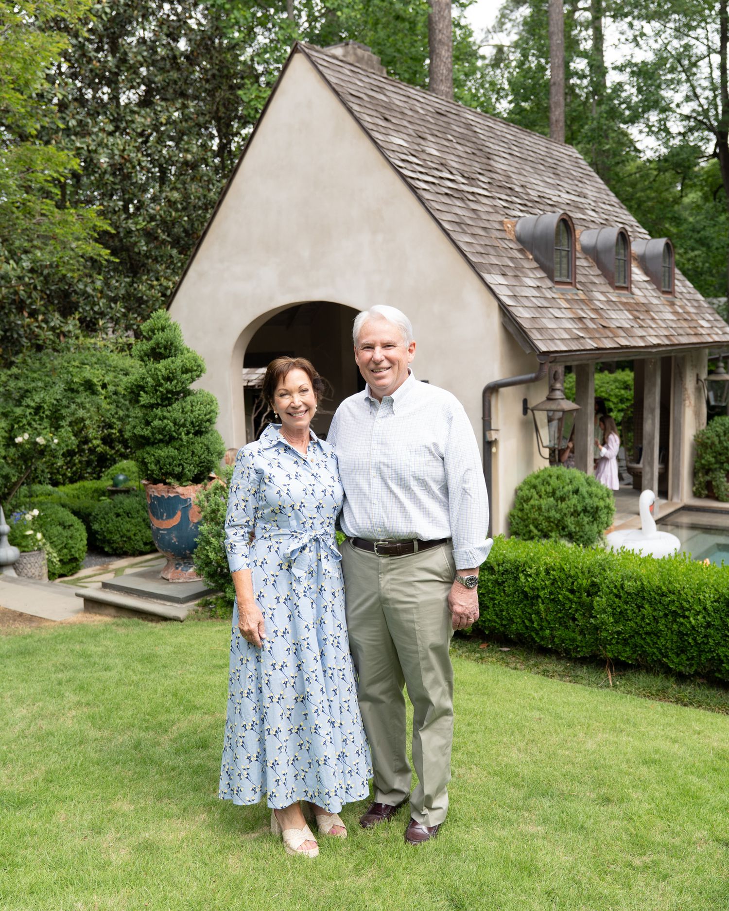 Smiling couple in garden attire poses in front of charming stucco cottage surrounded by manicured hedges.