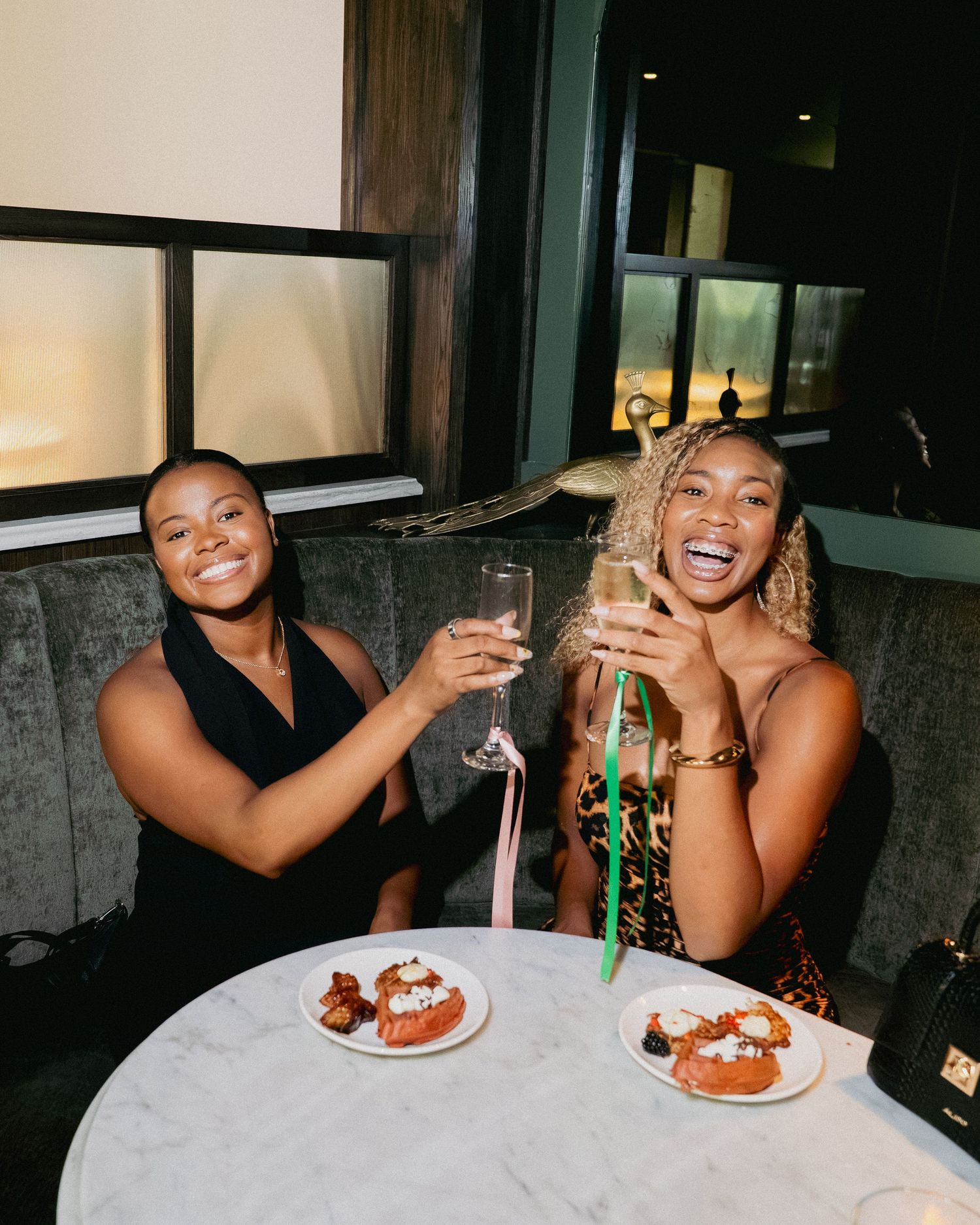 Two women smiling and toasting with champagne glasses at a restaurant table with desserts.