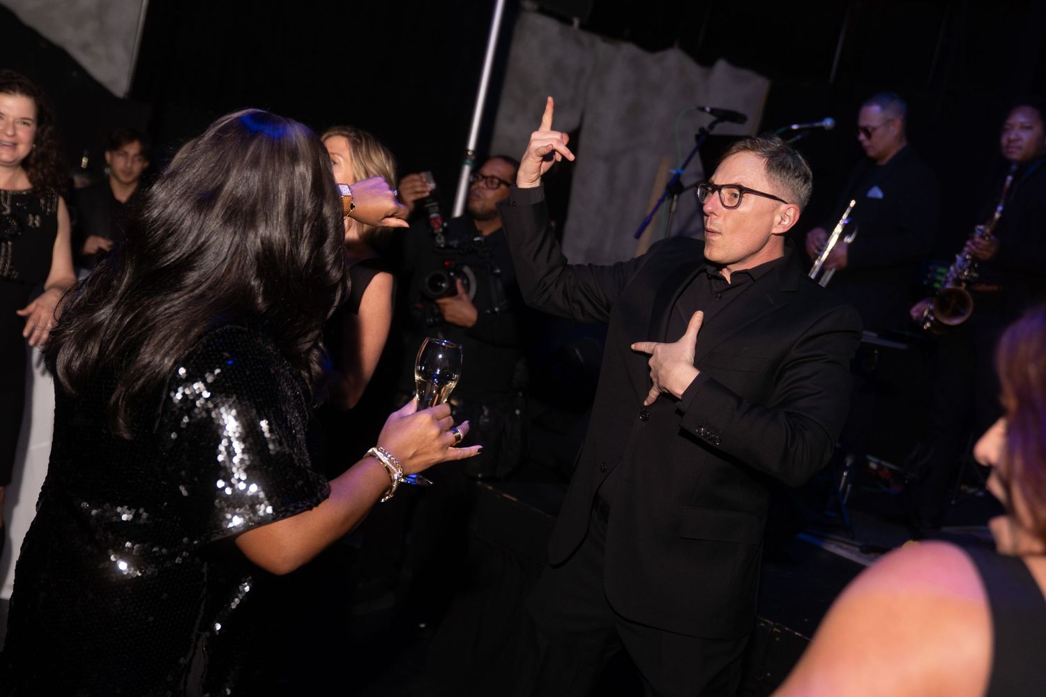 Man in black suit dancing and pointing up at an elegant evening party with live band performing in background.