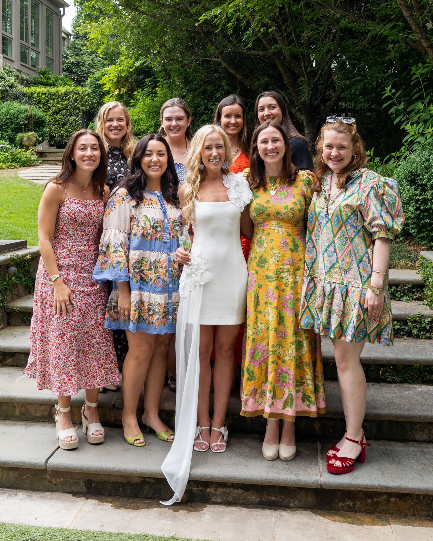 Bride in white dress poses with eight friends in colorful outfits on garden stone steps.