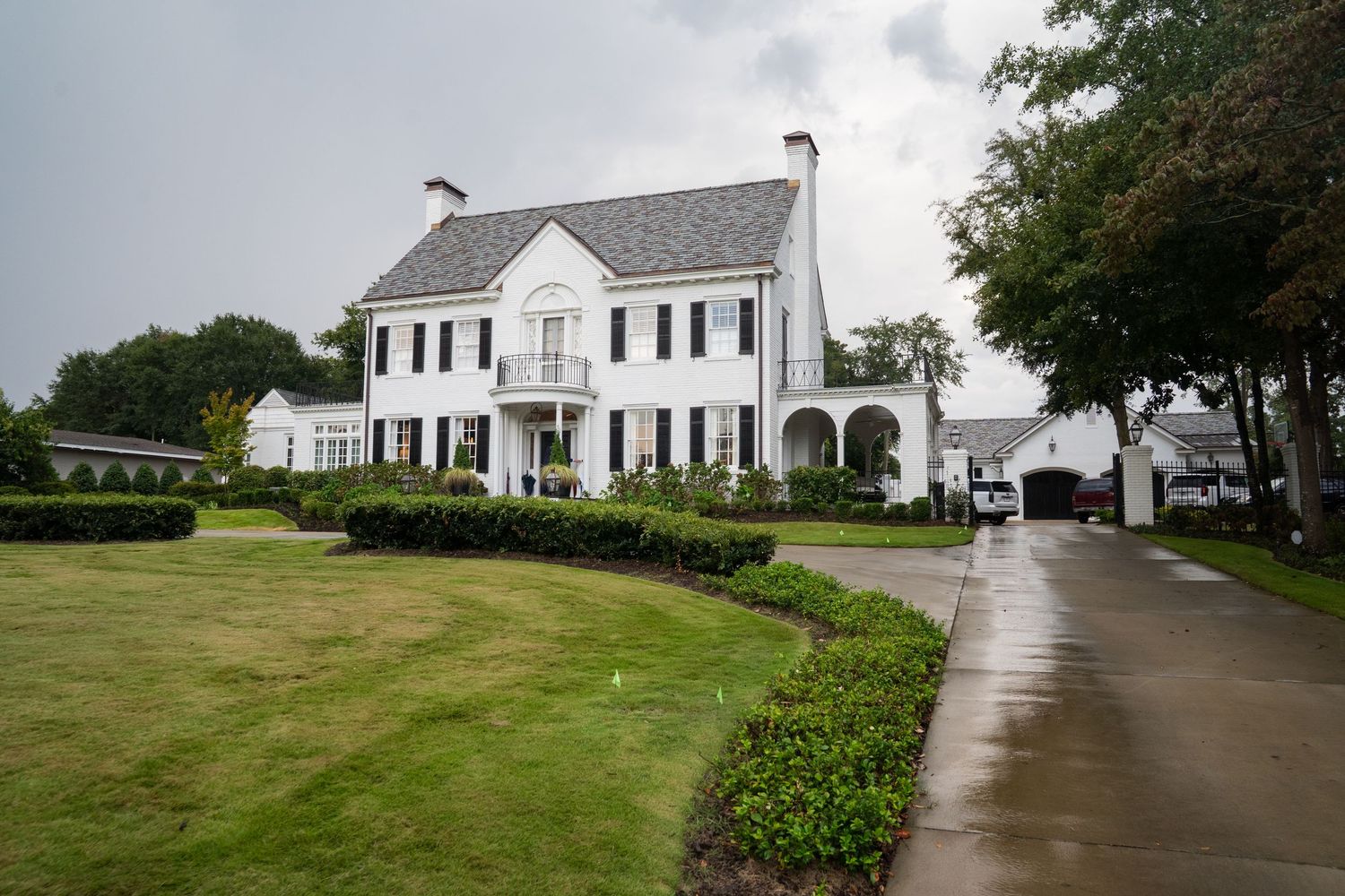 Elegant white colonial mansion with black shutters, manicured lawn, and curved driveway on a cloudy day.