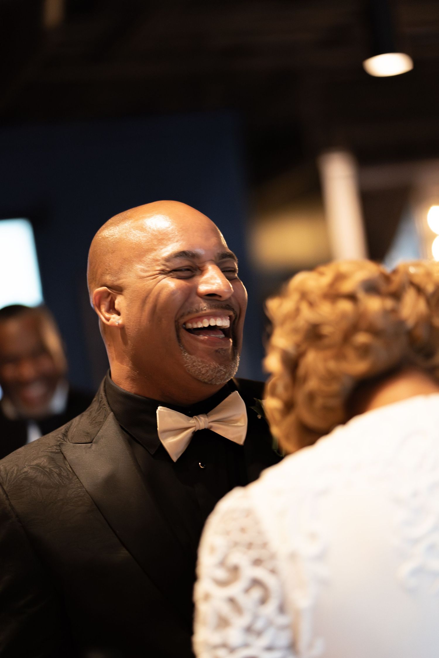 A bald man in a tuxedo laughs joyfully with a woman in white lace at a formal celebration.