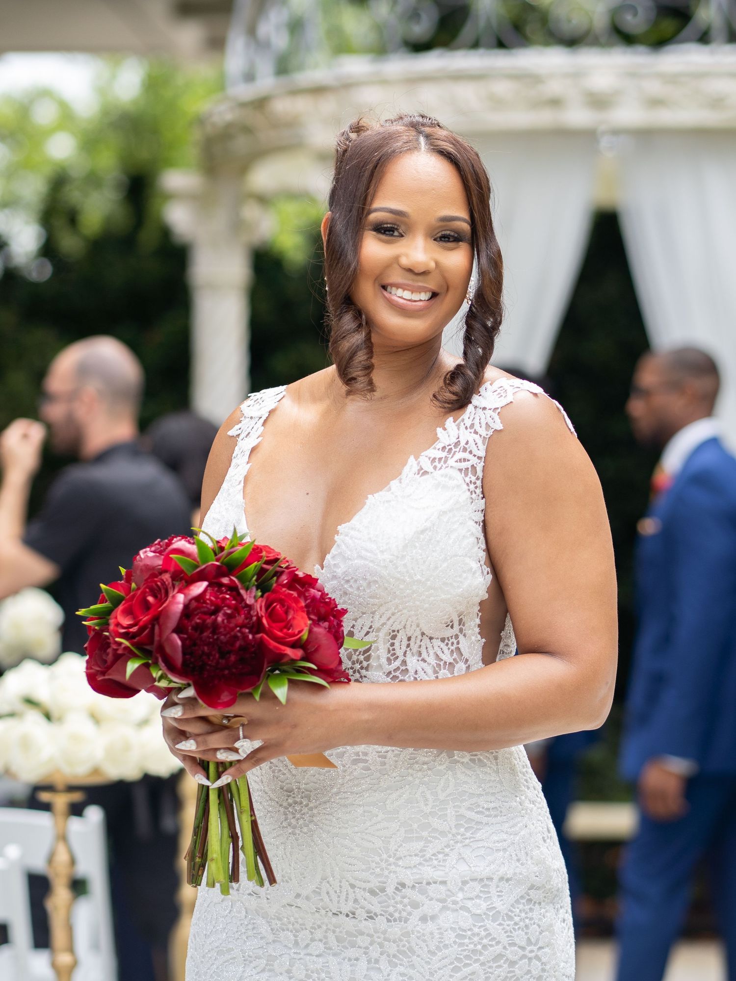 Bride in a beautiful lace wedding dress holding a vibrant red bouquet outdoors.
