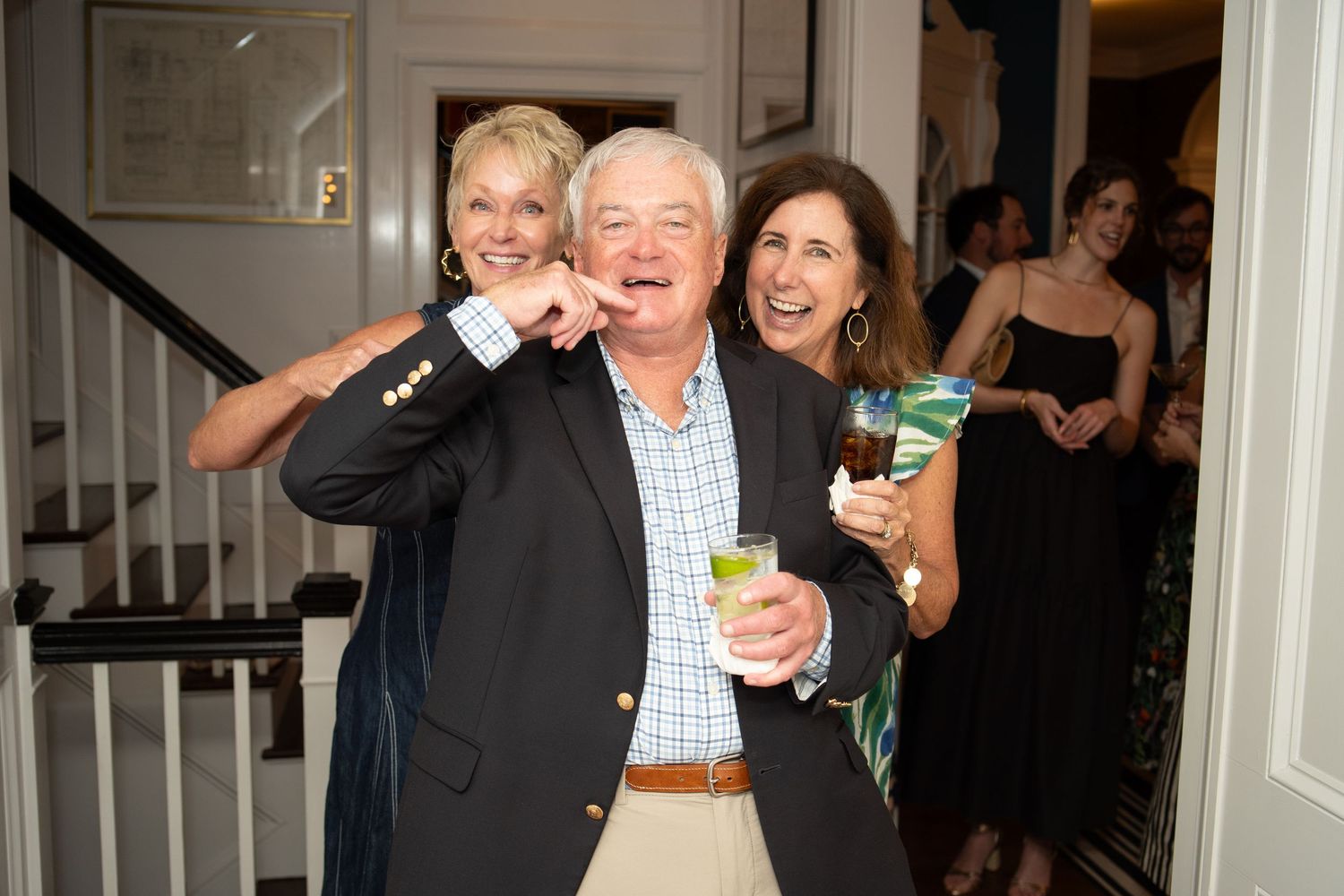 Man in navy blazer smiles holding a drink flanked by two laughing women at indoor party.