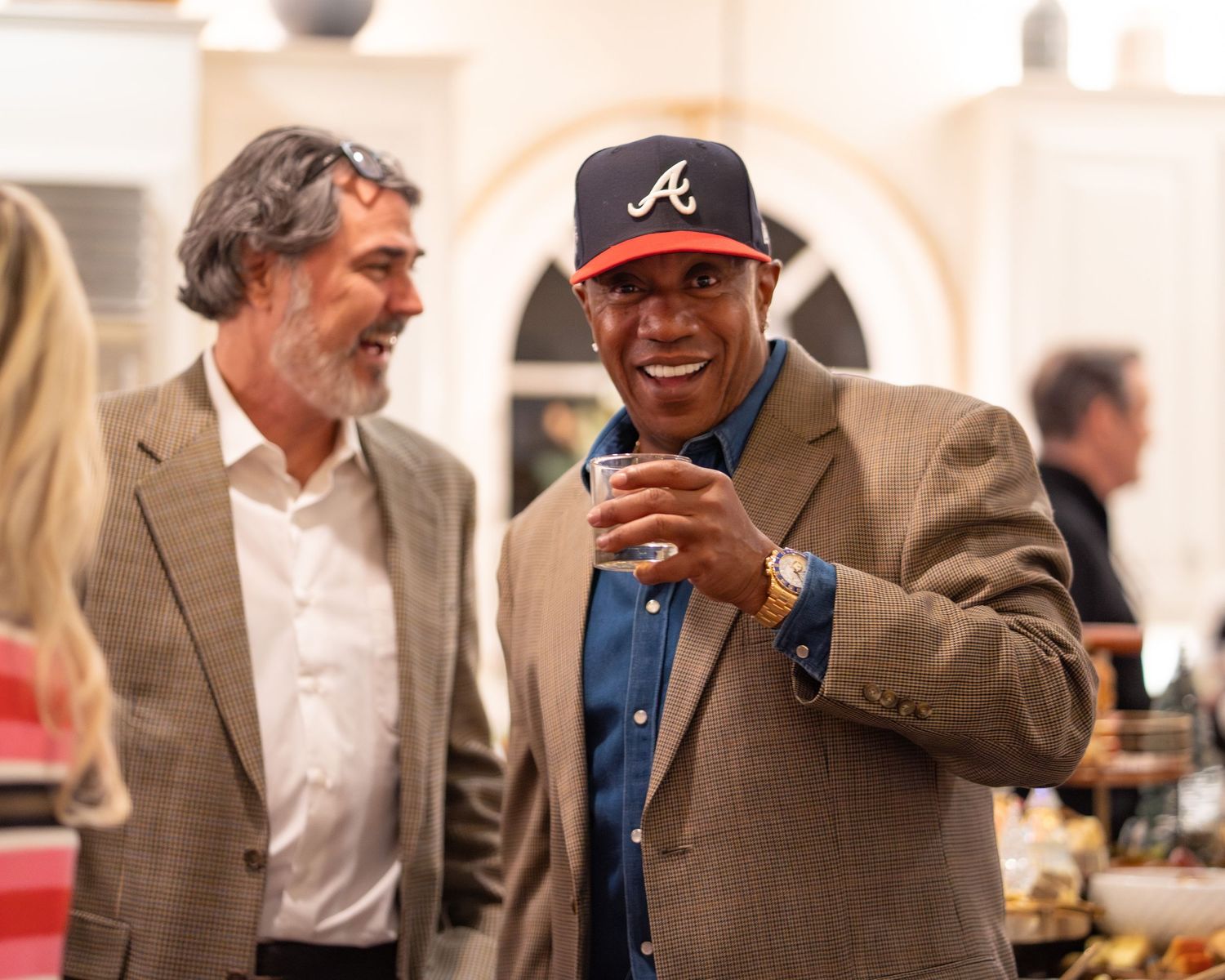 Two men laughing and socializing at a party, one wearing an Atlanta Braves cap and holding a drink.