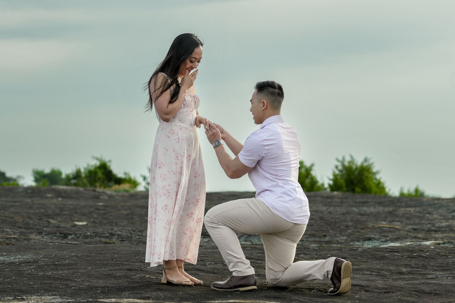 Man proposing on one knee to woman outdoors on rocky terrain with ring, romantic marriage proposal scene.